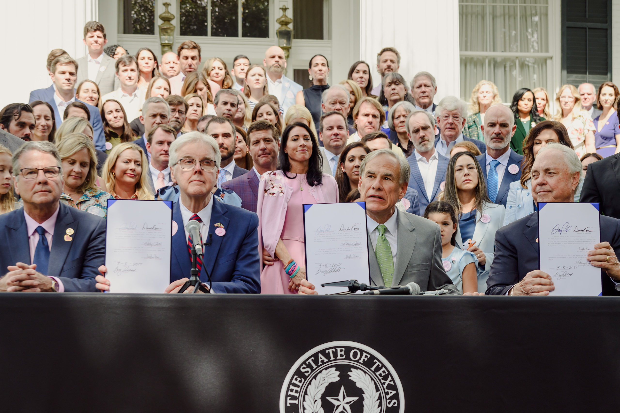 Gov. Greg Abbott, flanked by state Sen. Charles Perry, Lt. Gov. Dan Patrick and state Rep. Drew Darby, and backed by families of the Camp Mystic flood victims known as the “Heaven’s 27,” holds up the signed Senate Bill 1 and House Bill 1 at the Governor’s Mansion on Sept. 5, 2025. The bills, passed during the second special session of the 89th Texas Legislature, imposed stringent fees and requirements on camps.